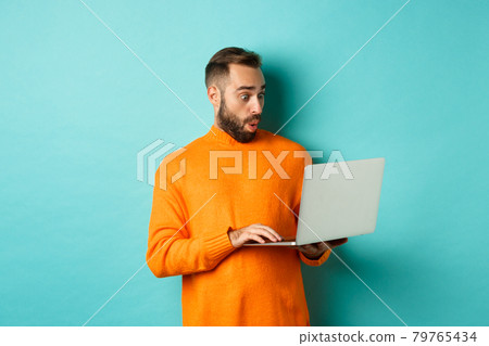 Freelance and technology concept. Image of handsome adult man staring at laptop with amazement, shopping online or working remote, standing over turquoise background 79765434