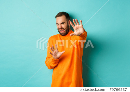Disgusted man cringe and extending hands in protection, begging for mercy, looking at something with aversion, standing over light blue background Disgusted man cringe and extending hands in protection, begging for mercy, looking at something with aversion, standing over light blue background 79766287