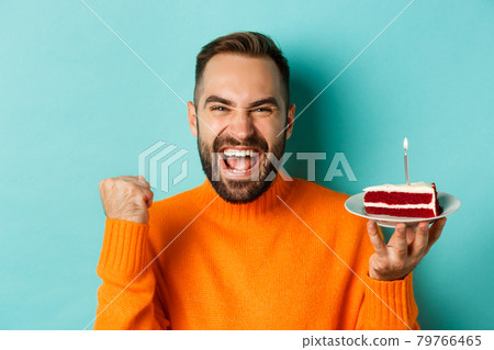 Close-up of happy adult man celebrating birthday, holding bday cake with candle and making wish, standing against turquoise background Close-up of happy adult man celebrating birthday, holding bday cake with candle and making wish, standing against turquoise background 79766465