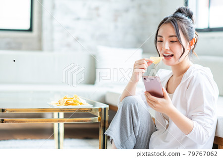 一位年輕女子一邊吃薯片一邊看著她的智能手機 一位年輕女子一邊吃薯片一邊看著她的智能手機 79767062