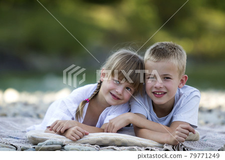 Two young happy cute blond smiling children, boy and girl, brother and sister laying embraced on pebbled beach on blurred bright sunny summer day background. Friendship and perfect holiday concept. Two young happy cute blond smiling children, boy and girl, brother and sister laying embraced on pebbled beach on blurred bright sunny summer day background. Friendship and perfect holiday concept. 79771249
