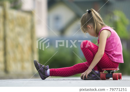 Pretty young long-haired blond child girl in casual pink clothing sitting on skateboard on paved suburb street on blurred sunny summer green background. Children activities, games and fun concept. Pretty young long-haired blond child girl in casual pink clothing sitting on skateboard on paved suburb street on blurred sunny summer green background. Children activities, games and fun concept. 79771251