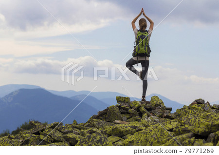 Back view of young slim tourist girl with backpack standing on one leg in yoga pose on rocky top on bright blue morning sky and foggy mountains background. Tourism, traveling and climbing concept. Back view of young slim tourist girl with backpack standing on one leg in yoga pose on rocky top on bright blue morning sky and foggy mountains background. Tourism, traveling and climbing concept. 79771269
