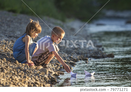 Two cute blond children, boy and girl on river bank sending in water white paper boats on bright summer blurred blue background. Joys and games of happy childhood and outdoors activities concept. Two cute blond children, boy and girl on river bank sending in water white paper boats on bright summer blurred blue background. Joys and games of happy childhood and outdoors activities concept. 79771271