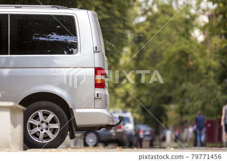 Side view detail of white passenger medium size luxury minibus van parked on summer city street pavement with blurred silhouettes of pedestrians on green trees bokeh copy space background. 79771456