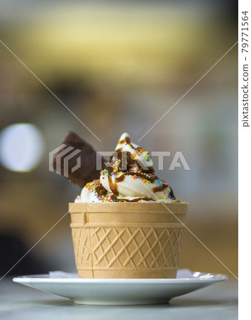 White plate with ice cream dessert in wafer cup with chocolate cookies and creative decoration topping on blurred colorful interior background. White plate with ice cream dessert in wafer cup with chocolate cookies and creative decoration topping on blurred colorful interior background. 79771564