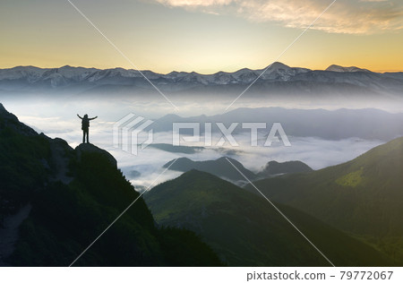 Wide mountain panorama. Small silhouette of tourist with backpack on rocky mountain slope with raised hands over valley covered with white puffy clouds. Beauty of nature, tourism and traveling concept 79772067