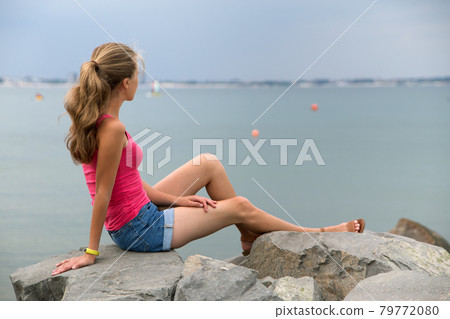 Young woman in summer clothes sitting on big boulders on sea shore looking on horizon. 79772080