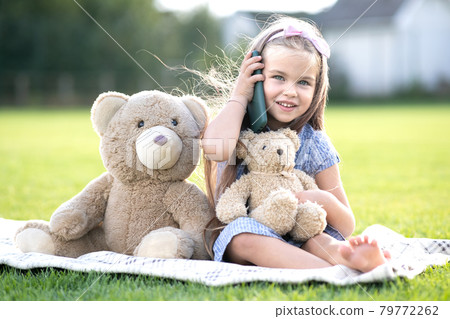 Pretty child girl sitting in summer park on green grass with her teddy bear toy talking on mobile phone outdoors in summer. 79772262