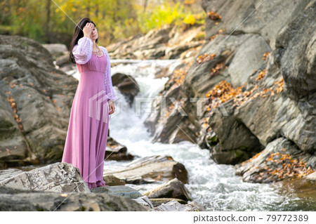 Young pretty woman in long fashionable dress standing near small mountain river with fast moving water. 79772389