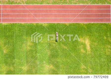 Aerial view of small figure of worker cutting green grass with mowing mashine on football stadium field with red running tracks in summer. 79772409
