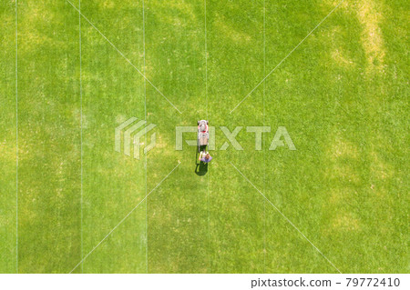 Aerial view of small figure of man worker trimming green grass with mowing mashine on football stadium field in summer. 79772410
