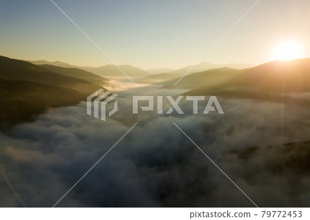 Aerial view of vibrant sunrise over white dense fog with distant dark silhouettes of mountain hills on horizon. 79772453