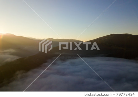 Aerial view of vibrant sunrise over white dense fog with distant dark silhouettes of mountain hills on horizon. 79772454