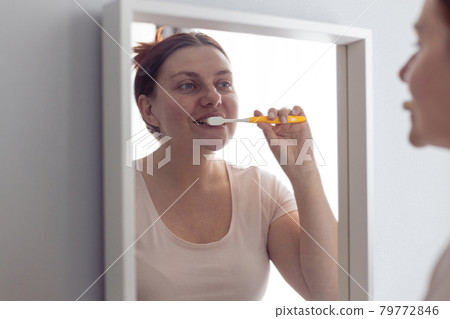 Portrait of beautiful millennial woman brushing teeth looking in mirror during morning hygiene procedures in the bathroom. Hygiene, dental care concept. 79772846