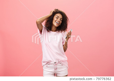 Portrait of carefree stylish modern dark-skinned young girl using smartphone hanging around joyfully touching hair and gazing with broad smile at camera holding cellphone posing against pink wall Portrait of carefree stylish modern dark-skinned young girl using smartphone hanging around joyfully touching hair and gazing with broad smile at camera holding cellphone posing against pink wall 79776362