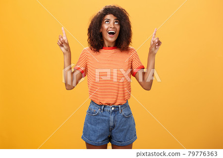 Fascinated impressed and amused good-looking charming African-American woman with afro hairstyle in trendy t-shirt and denim shorts pointing and looking up with interested look over orange wall 79776363