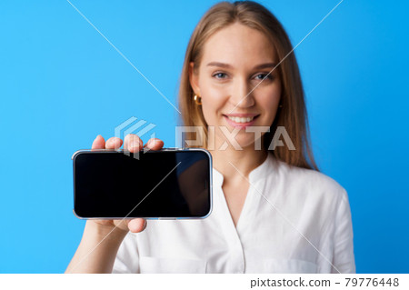 Beautiful young woman showing smartphone screen against blue background Beautiful young woman showing smartphone screen against blue background 79776448