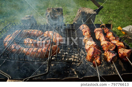 Barbeque meat and sausages or bratwurst on a grill grate in backyard. Man preparing shashlik or shish kebab over charcoal. Grilled meat on metal skewer outdoor. BBQ party or picnic food. Close up shot 79776613