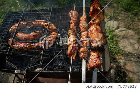 Barbeque meat and sausages or bratwurst on a grill grate in backyard. Man preparing shashlik or shish kebab over charcoal. Grilled meat on metal skewer outdoor. BBQ party or picnic food. Close up shot 79776623