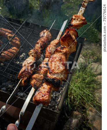 Barbeque meat and sausages or bratwurst on a grill grate in backyard. Man preparing shashlik or shish kebab over charcoal. Grilled meat on metal skewer outdoor. BBQ party or picnic food. Close up shot 79776624