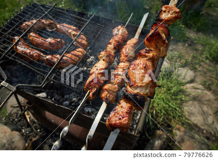Barbeque meat and sausages or bratwurst on a grill grate in backyard. Man preparing shashlik or shish kebab over charcoal. Grilled meat on metal skewer outdoor. BBQ party or picnic food. Close up shot 79776626