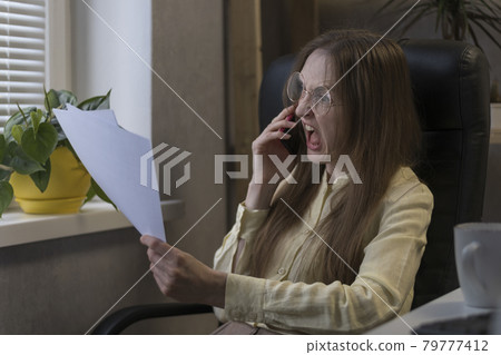 Angry woman boss talking on  phone and shouting, holding documents. Director scolds subordinates on smartphone. Angry woman boss talking on  phone and shouting, holding documents. Director scolds subordinates on smartphone. 79777412