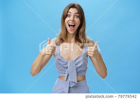 Woman satisfied with perfect result smiling excited with happy grin showing thumbs up gesture posing in stylish matching closed over blue background, staring thrilled and delighted at camera Woman satisfied with perfect result smiling excited with happy grin showing thumbs up gesture posing in stylish matching closed over blue background, staring thrilled and delighted at camera 79779542