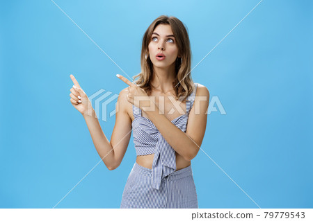 Woman getting interested in curious thing open mouth looking and pointing at upper left corner with intrigued focused expression being fascinated and surprised posing against blue background Woman getting interested in curious thing open mouth looking and pointing at upper left corner with intrigued focused expression being fascinated and surprised posing against blue background 79779543