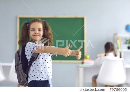 Adorable little girl with backpack showing thumbs up at classroom. Happy child going back to school, aiming for success 79786005