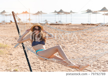 Enjoying summer vacation. Young happy woman wearing straw hat and sunglasses keeping eyes closed and relaxing while lying in the hammock on the sandy beach. People traveling concept Enjoying summer vacation. Young happy woman wearing straw hat and sunglasses keeping eyes closed and relaxing while lying in the hammock on the sandy beach. People traveling concept 79792789