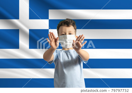 Little white boy in a protective mask on the background of the flag of Greece. Makes a stop sign with his hands, stay at home Greece 79797732