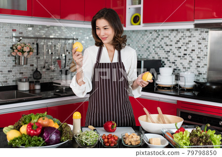 Beautiful middle age Asian woman housewife wearing apron standing new red tone kitchen and holding fresh lemon vegetable preparing vegetable salad Beautiful middle age Asian woman housewife wearing apron standing new red tone kitchen and holding fresh lemon vegetable preparing vegetable salad 79798850
