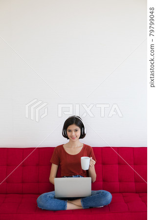 Half race Thai-German cute girl wearing headphone holding white coffee cup sitting on red sofa with business graph and chart and using laptop notebook computer work from home with happiness feeling 79799048