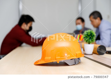 Close up image of a helmet on the desk with architects wearing protective masks working together in the backward, an architectural concept. 79800347