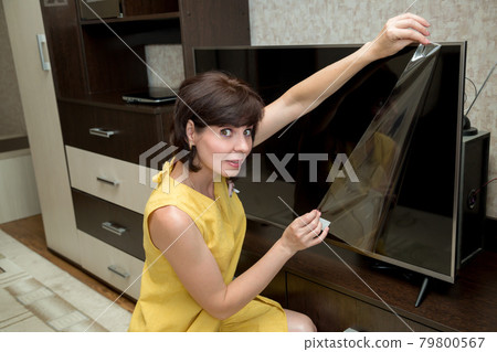 A brunette in a yellow dress in an apartment with a smile takes a film from a new large LCD TV A brunette in a yellow dress in an apartment with a smile takes a film from a new large LCD TV 79800567
