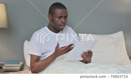 Young African Man Reacting to Loss on Documents in Bed Young African Man Reacting to Loss on Documents in Bed 79803710