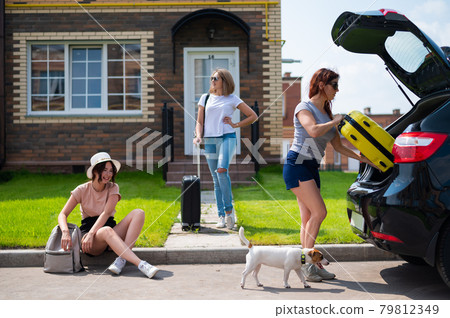 Three Caucasian women and a dog are going on a road trip. The girls are loading their suitcases into the trunk of the car. Summer vacation concept together with girlfriends. 79812349