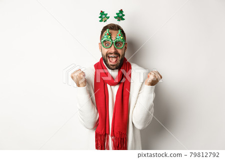 Christmas, New Year and celebration concept. Excited man winning prize, winter promotion, making fist pump and celebrating, wearing party glasses, white background 79812792