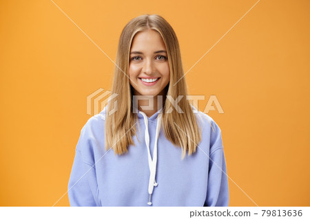 Waist-up shot of pleasant kind and happy friendly-looking young fair-haired girl in comfortable hoodie smiling cheerfully at camera with delighted carefree look posing against orange background Waist-up shot of pleasant kind and happy friendly-looking young fair-haired girl in comfortable hoodie smiling cheerfully at camera with delighted carefree look posing against orange background 79813636