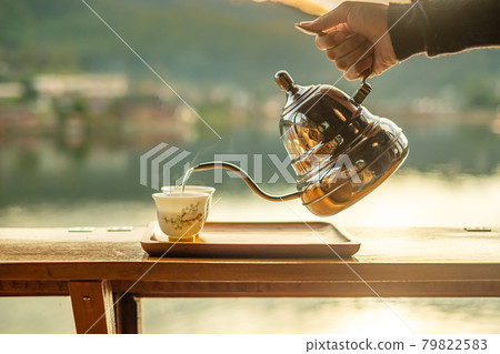 Hand holding vintage teapot and pouring hot tea to cup on wood table against lake view background at coffee shop in the morning sunrise, Ban Rak Thai village, Mae Hong Son province, Thailand Hand holding vintage teapot and pouring hot tea to cup on wood table against lake view background at coffee shop in the morning sunrise, Ban Rak Thai village, Mae Hong Son province, Thailand 79822583