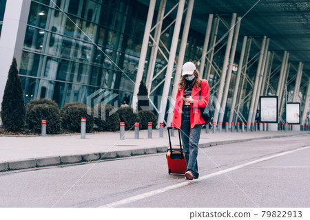 Portrait of a traveler woman in a mask walking with an orange suitcase near an airport. Young fashionable woman in a blue jeans and jacket 79822913
