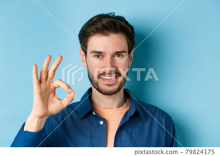 Close up of handsome bearded guy showing okay gesture and smiling, standing on blue background Close up of handsome bearded guy showing okay gesture and smiling, standing on blue background 79824175