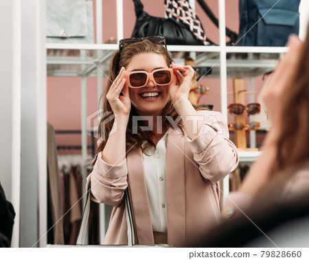 Laughing woman try on sunglasses in front of a mirror in a fashion store Laughing woman try on sunglasses in front of a mirror in a fashion store 79828660