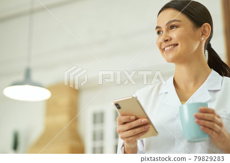 Happy female doctor standing with smartphone and cup 79829283