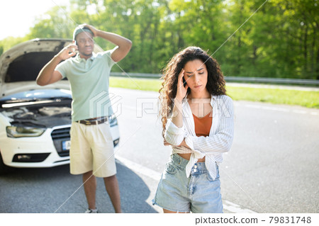 Young stressed woman standing on highway near broken car while boyfriend calling breakdown road service on smartphone Young stressed woman standing on highway near broken car while boyfriend calling breakdown road service on smartphone 79831748