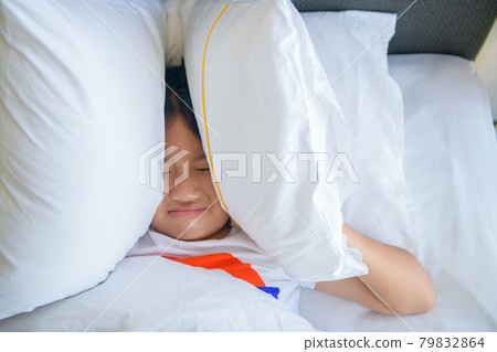Little girl lying in bed covering head with pillow because too loud annoying noise Little girl lying in bed covering head with pillow because too loud annoying noise 79832864