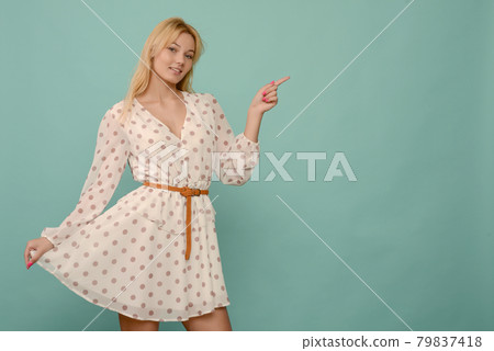 Delighted beautiful young woman in white dress in polka dots posing in studio on blue background. 79837418