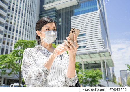 A three-quarter sleeve business woman operating a smartphone while wearing a mask in the business district in front of Nagoya Station 79843522
