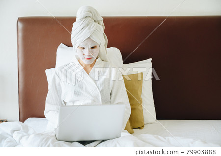 Portrait of young woman applying facial mask on her face during using computer laptop on bedroom. Conceptual of young woman lifestyle during working from home. Portrait of young woman applying facial mask on her face during using computer laptop on bedroom. Conceptual of young woman lifestyle during working from home. 79903888